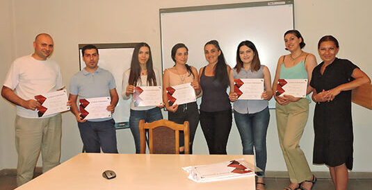 young adults holding TEFL certificates stand with their instructor in front of a decorative stone wall carving—a cozy scene celebrating the completion of a 120‑hour TEFL training.